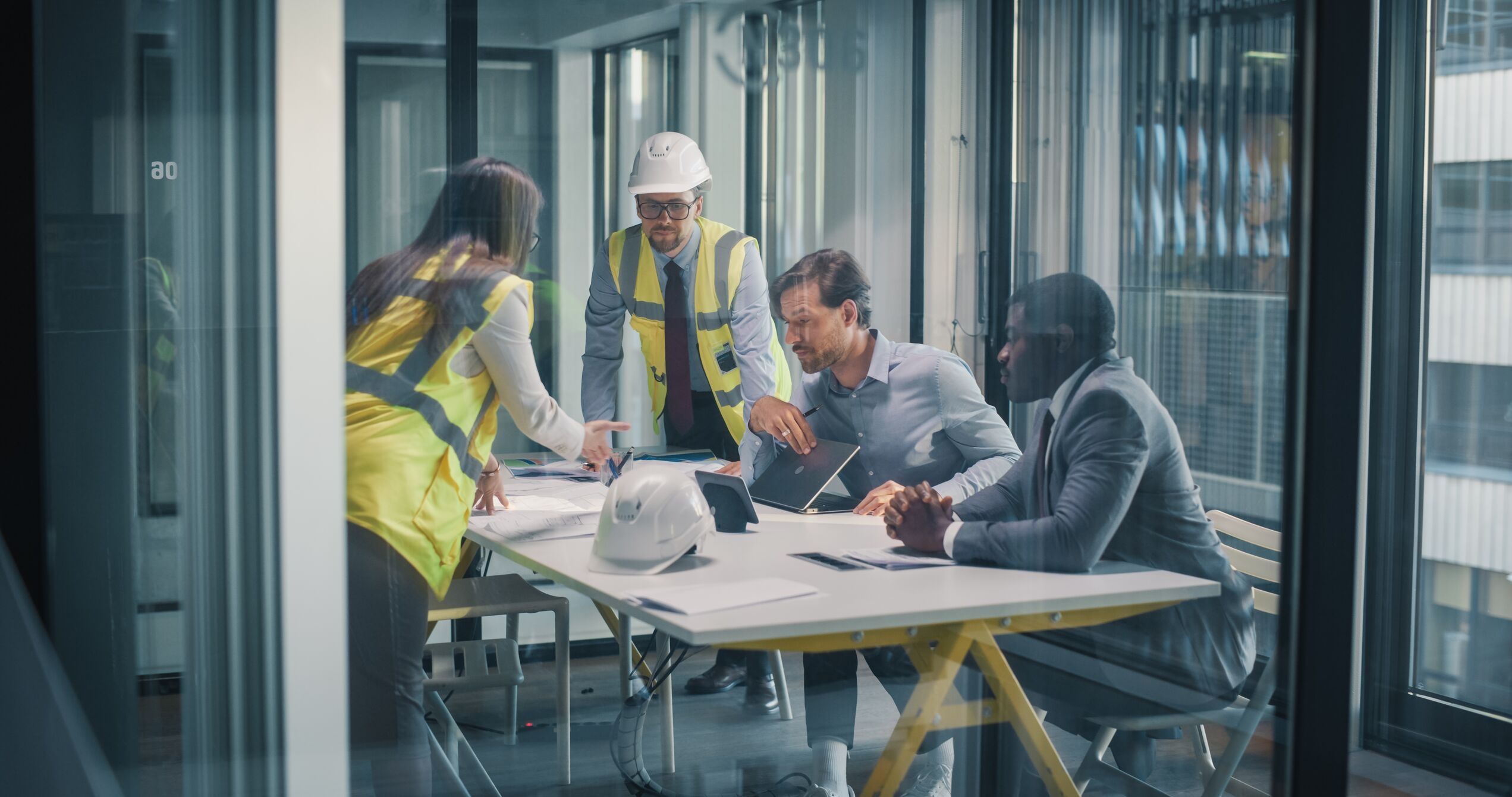 Four people, two in construction vests and hard hats, other in business attire around a table planning